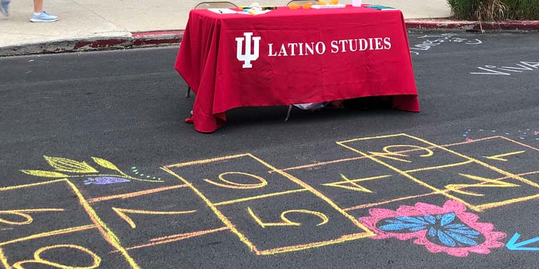 A table with a red tablecloth and Latino Studies sign behind a colorful chalk hopscotch drawing.